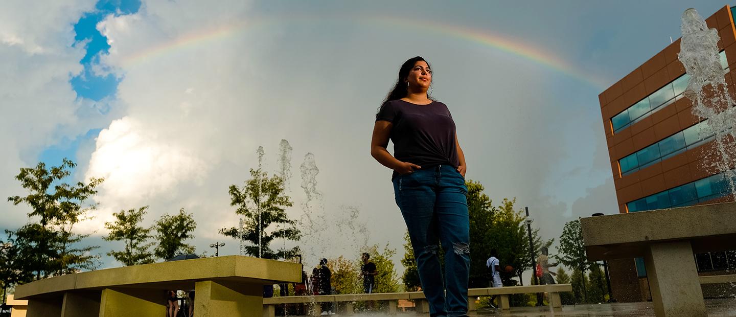 A young woman standing with her hands in her pockets on Oakland University's campus, with a rainbow and fountain behind her.